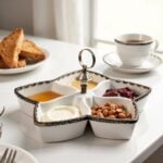 Ceramic divided plate with silver accents serving breakfast items, honey, jam, and nuts