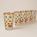 Line of six hand-painted Moroccan tea glasses with gold rims and bright floral motifs on neutral background.