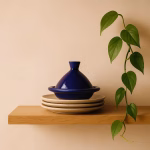Royal-blue mini tagine displayed on stacked neutral stoneware plates atop a wooden shelf with a trailing plant.