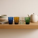 Shelf display of blue, amber, clear, and green glass tumblers beside ceramic teapot.