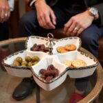 Hand holding white ceramic divided plate with silver accents filled with olives, dates, and crackers