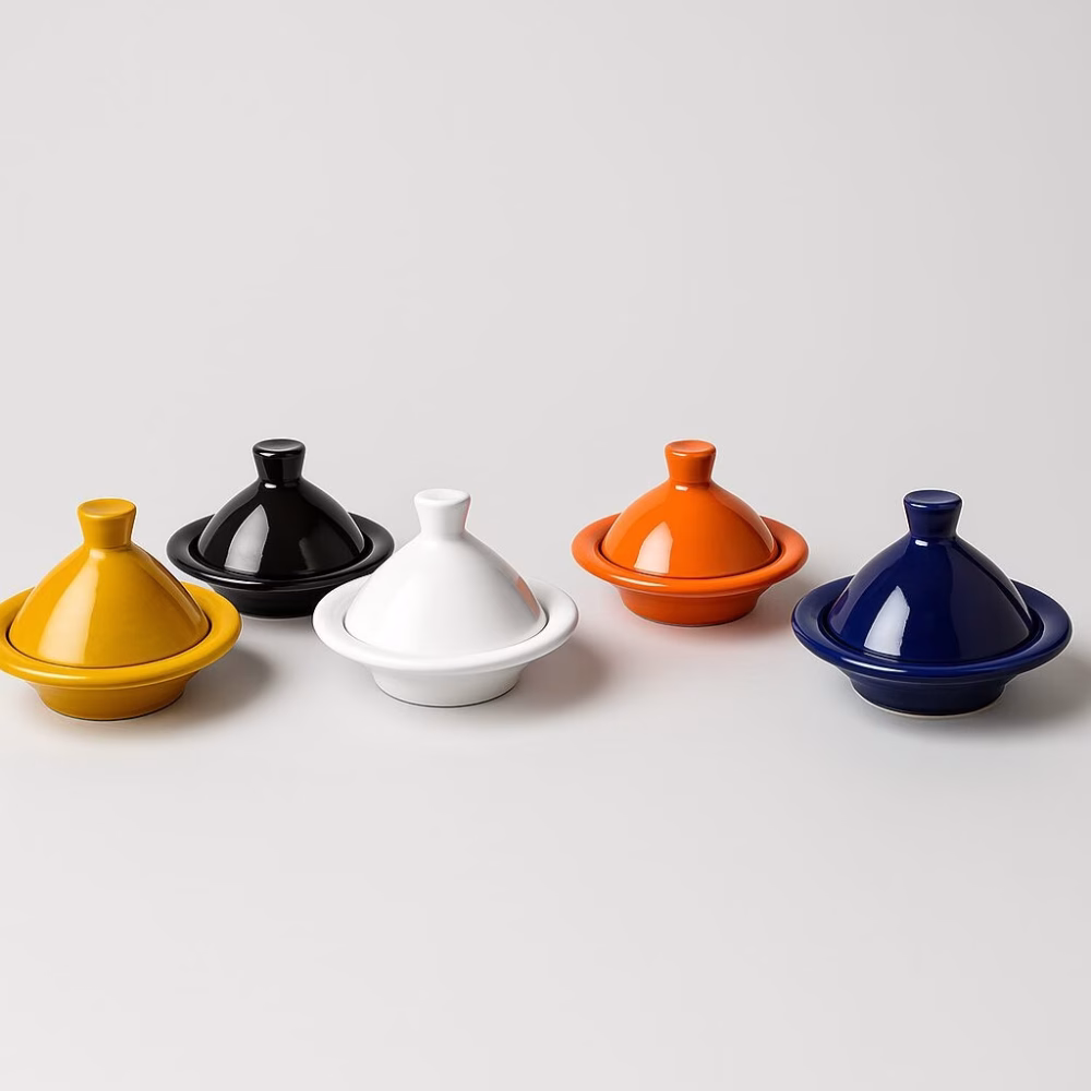 Studio lineup of five glazed mini tagines—mustard, black, white, orange, and royal blue—arranged in a row against a light-grey background.