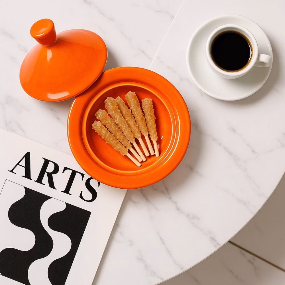 Top-down view of an orange mini tagine lid and base holding rock-candy sticks next to a black-coffee cup and modern art magazine on a white marble surface.