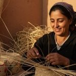 Smiling Moroccan straw artisan weaving raffia basket in sunlit workshop, straw stacks behind her.