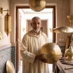 Master Moroccan brass lamp artisan Abdul Jalil holding a polished copper lantern in his well-lit Souq Nhas workshop, Marrakesh.