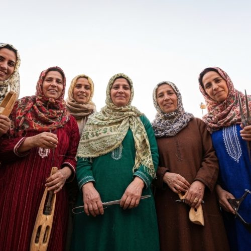 A low-angle group portrait of seven Beni Ourain rug weavers, smiling and holding their weaving tools, including shuttles and combs.