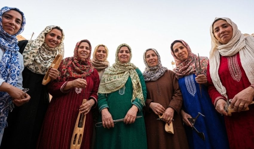 A low-angle group portrait of seven Beni Ourain rug weavers, smiling and holding their weaving tools, including shuttles and combs.