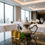 A hand-engraved traditional silver teapot resting on a dark marble table in an elegant, modern restaurant setting, served alongside two glasses of fresh Moroccan mint tea.