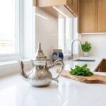 A hand-engraved traditional silver teapot displayed on a bright white quartz countertop in a modern Scandinavian-style kitchen, styled next to a rustic wooden board with fresh mint leaves.