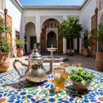 A hand-engraved traditional silver teapot beautifully presented on a vibrant geometric zellige tile table in a sunlit Moroccan courtyard, accompanied by classic gold-rimmed tea glasses and fresh mint.
