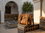 Vintage leather satchel bag displayed on mosaic tile pedestal in Moroccan courtyard with fountain and orange tree