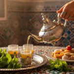 authentic Moroccan tea ceremony scene featuring your beautiful hand-engraved silver teapot with the ornate tripod feet. The image shows it pouring tea into ornate cups on a hammered silver tray, surrounded by fresh mint sprigs and Moroccan sweets, with a hand wearing traditional jewelry.