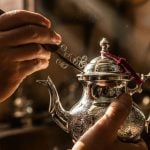 Close-up of a Maalem artisan's hands hand-engraving an intricate floral pattern onto a silver Moroccan teapot using the naqsh technique.