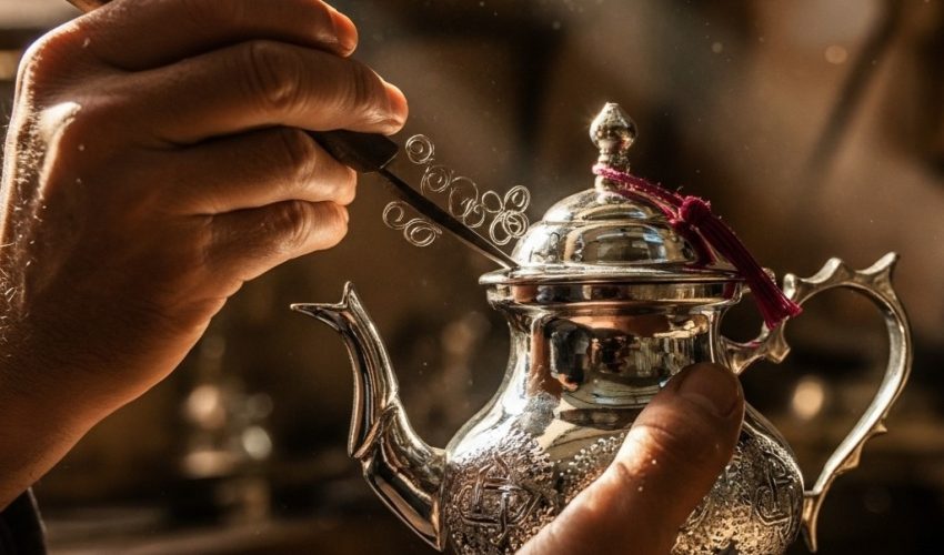 Close-up of a Maalem artisan's hands hand-engraving an intricate floral pattern onto a silver Moroccan teapot using the naqsh technique.