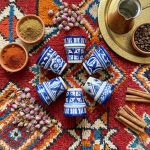 Six handpainted ceramic espresso cups arranged in circle on colorful Berber rug with spices, rose buds and coffee pot