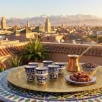 Ceramic espresso cups on brass tray with dates and coffee pot, rooftop view of Marrakech medina and Atlas Mountains
