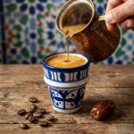 Hot espresso being poured from copper cezve into handpainted ceramic cup with coffee beans and date on wooden table