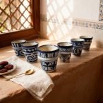 Six ceramic espresso cups on terracotta windowsill with traditional mashrabiya screen, dates and brass spoon