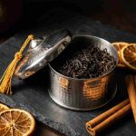 Silver hammered metal box with tea leaves, dried orange slices, and cinnamon on dark slate