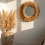 Natural raffia round mirror with plush shaggy straw fringe above ceramic vase with pampas grass in minimalist interior