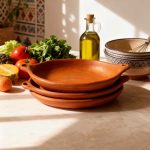 Stack of three terracotta baking dishes on kitchen counter with fresh vegetables and olive oil