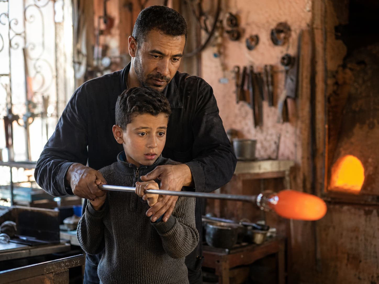 Younes guides his son's hands on the kasaba blowpipe, passing down three generations of Moroccan glassblowing knowledge
