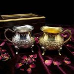 Hand-engraved silver and brass mini creamer pitchers displayed on burgundy velvet with dried rose petals