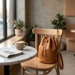 Tan leather bucket bag on wooden chair in minimalist café setting