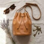 Tan leather bucket bag flat lay top view with sunglasses lavender and keys