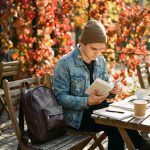 Man reading at outdoor café with leather daypack on chair beside him
