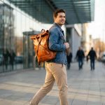 Man wearing tan leather roll top backpack walking in modern city environment