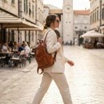 Woman wearing a minimalist leather backpack in cognac brown while walking through a European cobblestone plaza, styled with a cream linen blazer