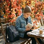 Navy blue minimalist leather backpack resting on a wooden café chair beside a man reading during autumn, perfect for everyday carry