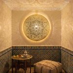 Round brass wall sconce casting radiant shadow patterns in traditional room with colorful zellige tile walls leather pouf and brass tea service
