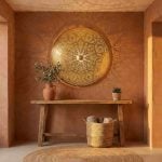Brass wall sconce illuminating warm terracotta entryway with rustic wooden bench olive branches and woven basket casting sunburst shadows