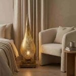 Hand-pierced brass teardrop floor lamp glowing warmly between bed and boucle armchair in neutral toned bedroom with linen curtains