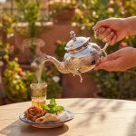 Hand pouring steaming mint tea from silver teapot with diamond pattern into traditional glass with Moroccan pastries