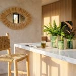 A Handwoven Palm Leaf Bar Chair sits at a sunlit kitchen island with green Tamegroute plant pots and a straw eye mirror on the wall.