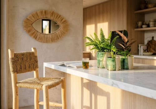 A Handwoven Palm Leaf Bar Chair sits at a sunlit kitchen island with green Tamegroute plant pots and a straw eye mirror on the wall.
