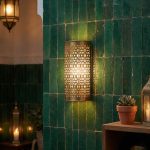 Moroccan brass filigree wall light glowing against traditional green zellige tiled wall in riad-style interior