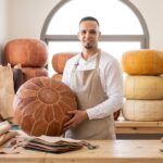 Marrakech leather artisan Abdelilah holding a finished handmade leather pouf in his bright workshop at Souk Tala'a