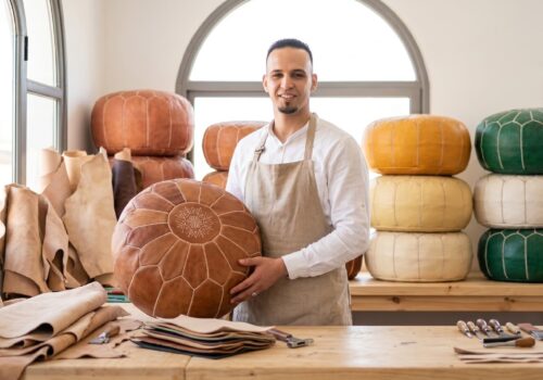 Marrakech leather artisan Abdelilah holding a finished handmade leather pouf in his bright workshop at Souk Tala'a