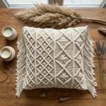 Handwoven macrame cushion cover with diamond knotwork pattern and fringe displayed on wooden table with candles and dried lavender