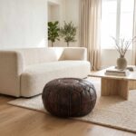 Brown leather pouf on a wool rug between a boucle sofa and a travertine table