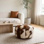 Brown and white cowhide pouf on a wool rug beside a boucle sofa and travertine coffee table