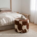 Brown and white cowhide ottoman used as a bedside table with books and a mug