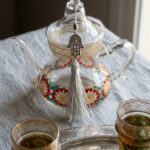 Close-up of borosilicate glass teapot with gold lattice band, hand-painted rosettes and silver Hamsa tassel