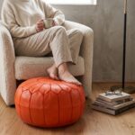 Orange leather pouf used as a footrest in front of a cream boucle armchair
