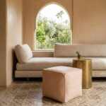 Undyed leather cube pouf beside a linen sofa and hammered brass side table under an arched window